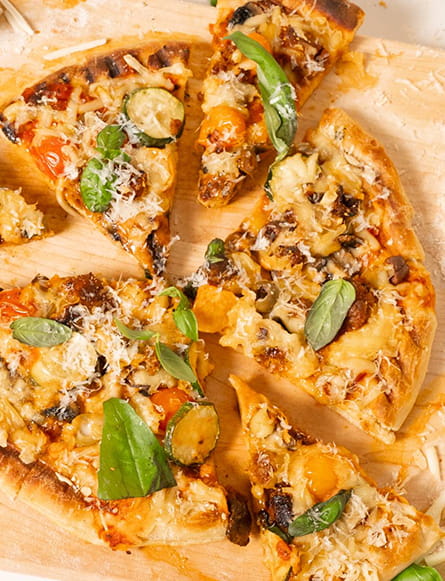 A sliced grilled vegetable pizza topped with melted cheese, zucchini, tomatoes, mushrooms and fresh basil on a wooden board.