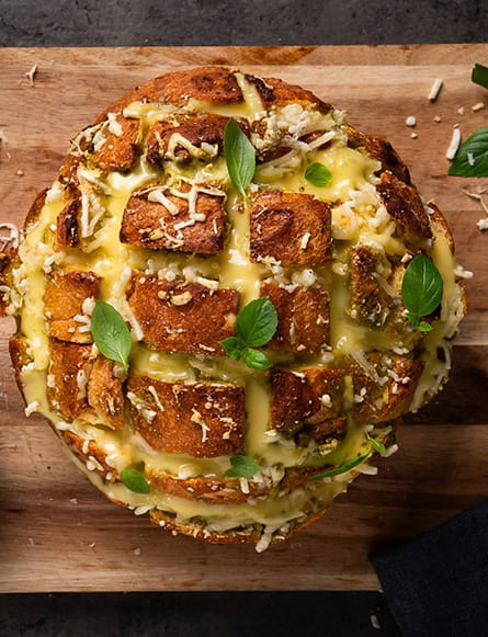 A loaf of hedgehog‑style garlic bread scored into cubes, filled with melted cheese and topped with herbs, placed on a wooden board.