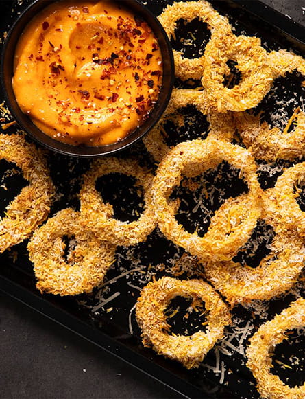 A tray of crispy baked onion rings coated in breadcrumbs, served with a bowl of creamy dipping sauce topped with chili flakes.