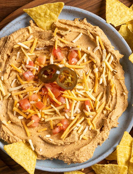 A plate of creamy vegan taco dip topped with dairy free shreds, diced tomatoes and sliced jalapeños, surrounded by tortilla chips.