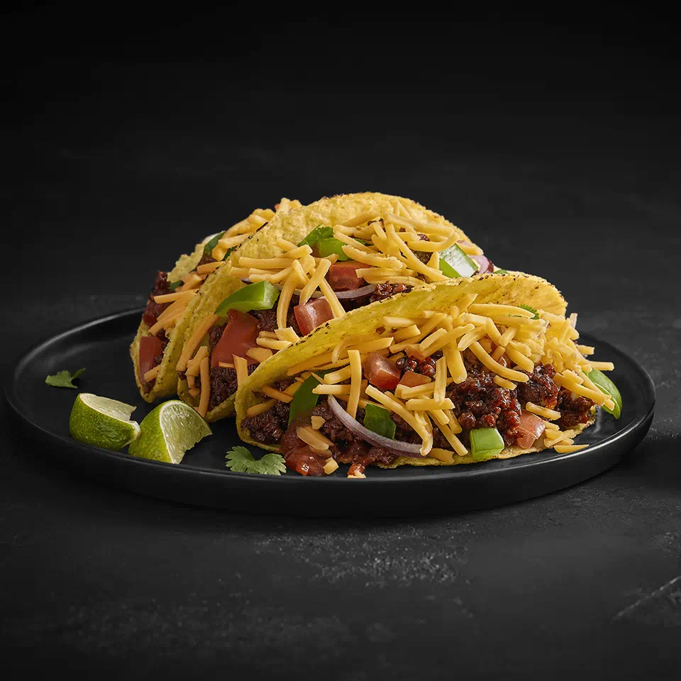 A plate with two tacos filled with dairy free cheese shreds, diced vegetables and garnished with lime wedges, set against a dark background.