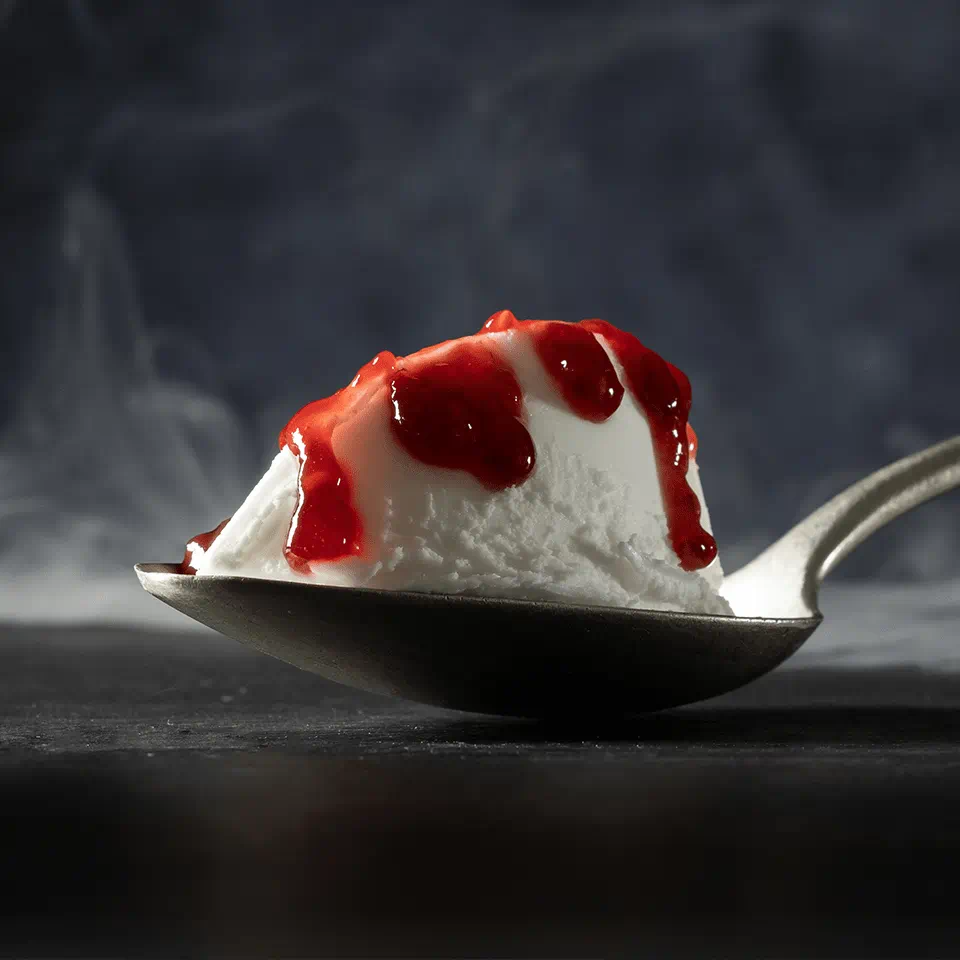 A metal spoon holds a small scoop of dairy free cream cheese drizzled with strawberry sauce, with a dark, smoky background.