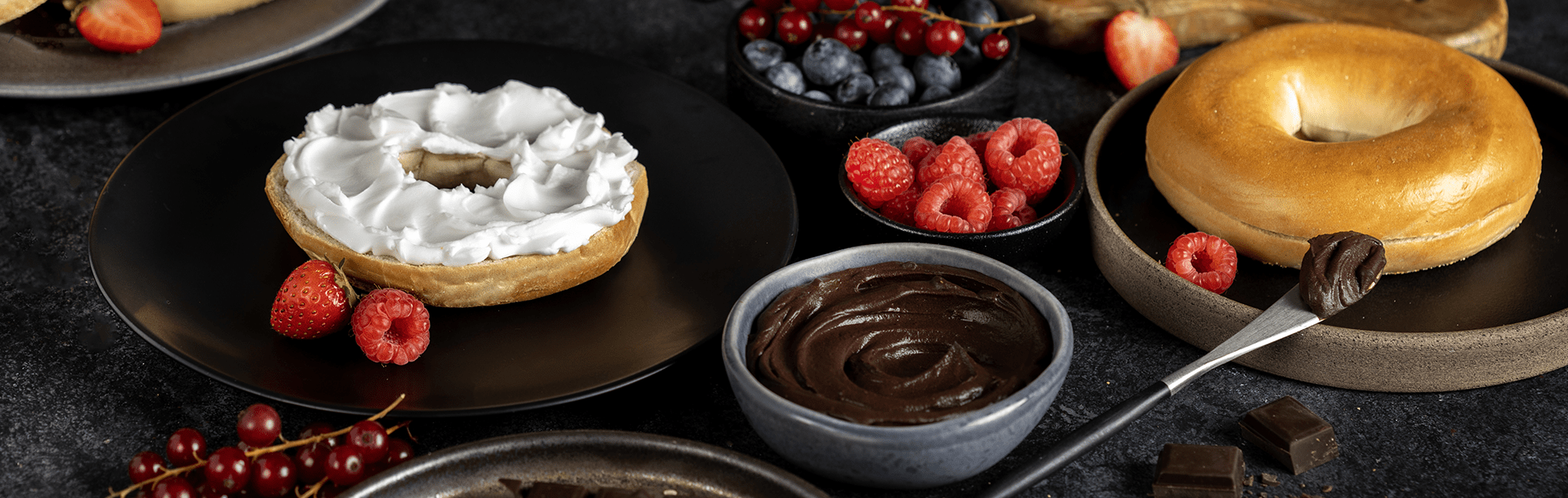 A cream cheese covered half bagel and a plain bagel sit on dark plates among bowls of mixed berries and chocolate spread.
