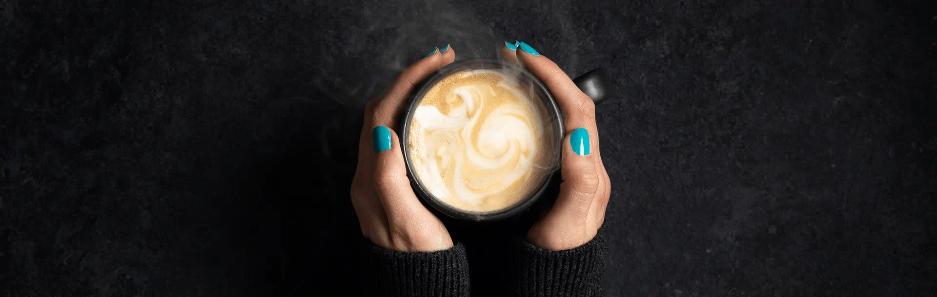 A person with turquoise nail polish holds a warm latte in a black mug, viewed from above, with a smooth swirl in the foam.