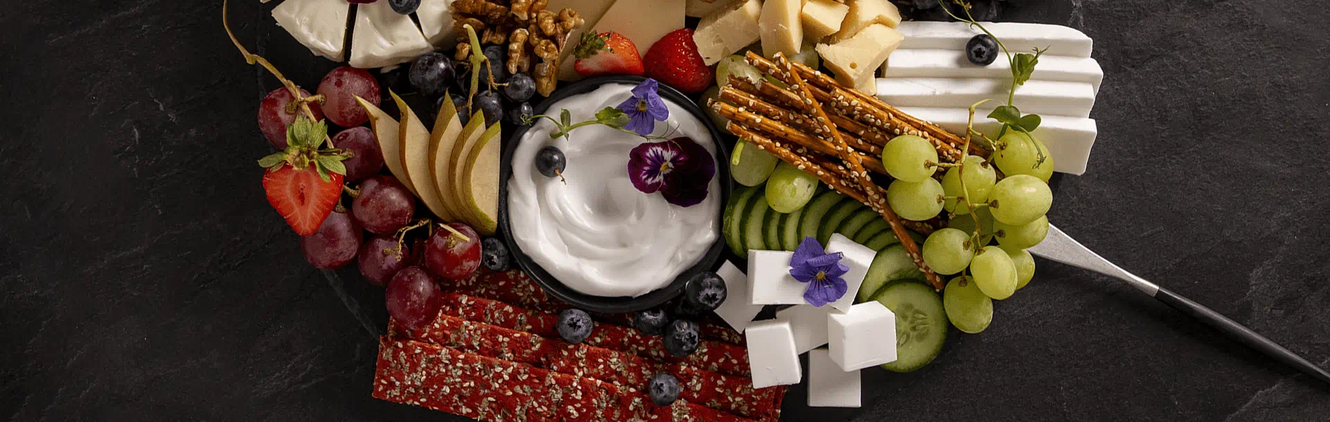 A grazing board with mixed fruits, pretzels, nuts, seed crackers, dairy free cheese and a bowl of dip topped with blueberries and an edible flowers.