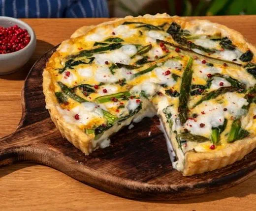Baked asparagus quiche filled with Violife dairy free products, green asparagus and pink peppercorns served on a round wooden board.