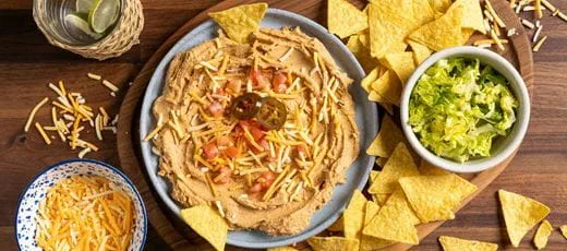 Vegan taco dip topped with dairy free cheese shreds, diced tomatoes and sliced jalapeños, surrounded by yellow corn tortilla chips.