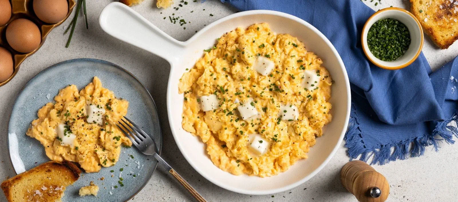 Creamy scrambled eggs in a skillet, accompanied by toasted bread slices. Dish is toped with chopped chives and cubes of Violife plant butter.