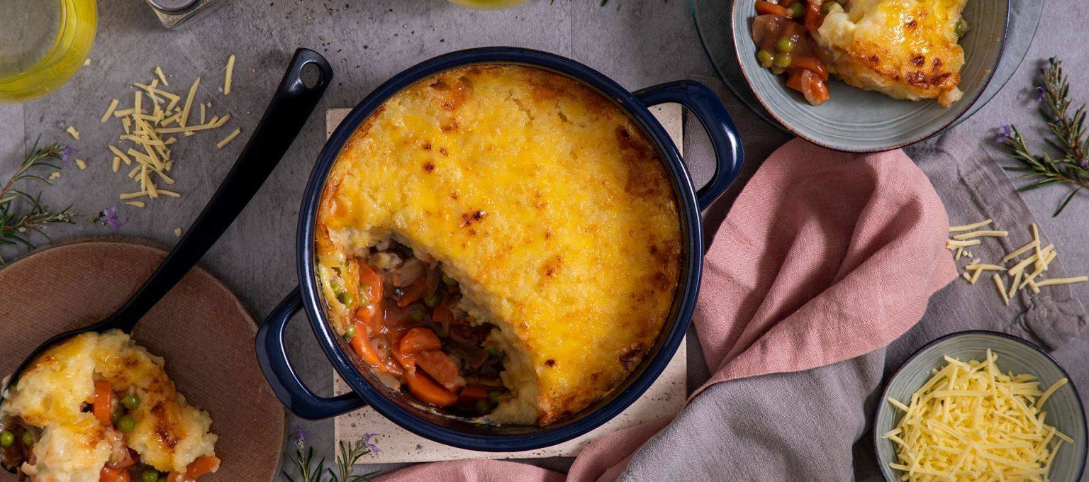 Vegan shepherd’s pie in a round baking dish, filling made with carrots, peas, and mushrooms with a golden-brown mashed topping of melted dairy free cheese. 
