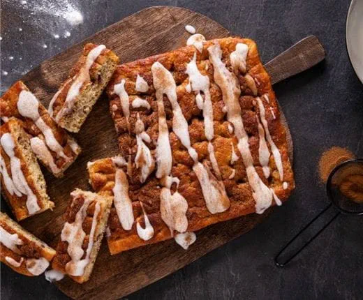 A cinnamon roll focaccia, on a wooden serving board, topped with swirls of cinnamon sugar, drizzled with dairy free cream cheese frosting.