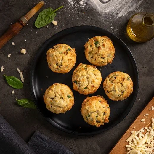 A black plate holding six spinach muffins with visible green spinach pieces. Background is dark rustic surface sprinkled with flour and dairy free shreds.