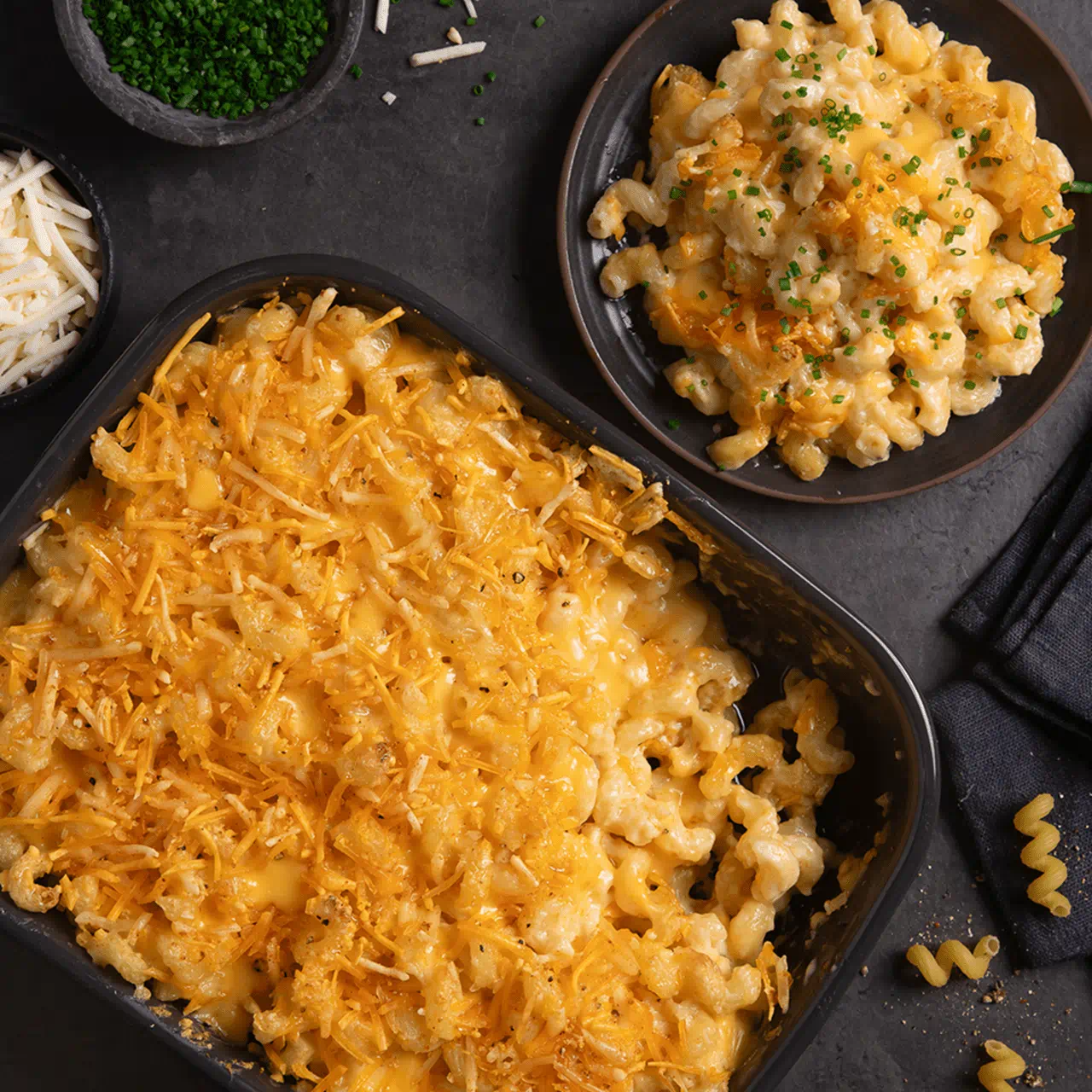 A baking dish filled with golden, cheesy homemade mac and cheese made with cavatappi pasta. Bowls of dairy free cheese shreds and  chives are placed nearby.