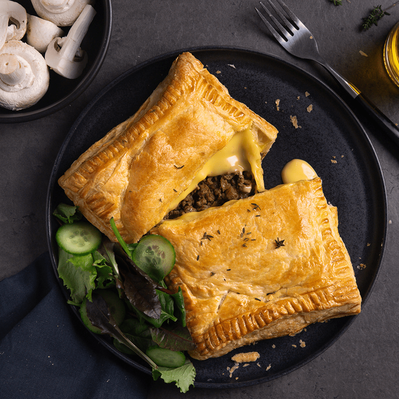 A puff pastry pocket sits on a dark plate, split open to reveal a melted cheesy filling. Fresh green salad and sliced cucumbers are arranged beside it.