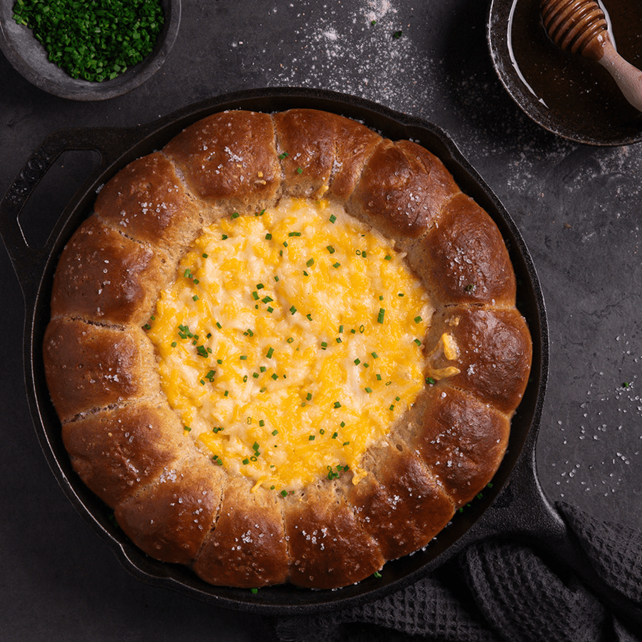 A cast‑iron skillet filled with a ring of golden‑brown pretzel bites surrounding a creamy, beer cheesy dip topped with chopped chives.
