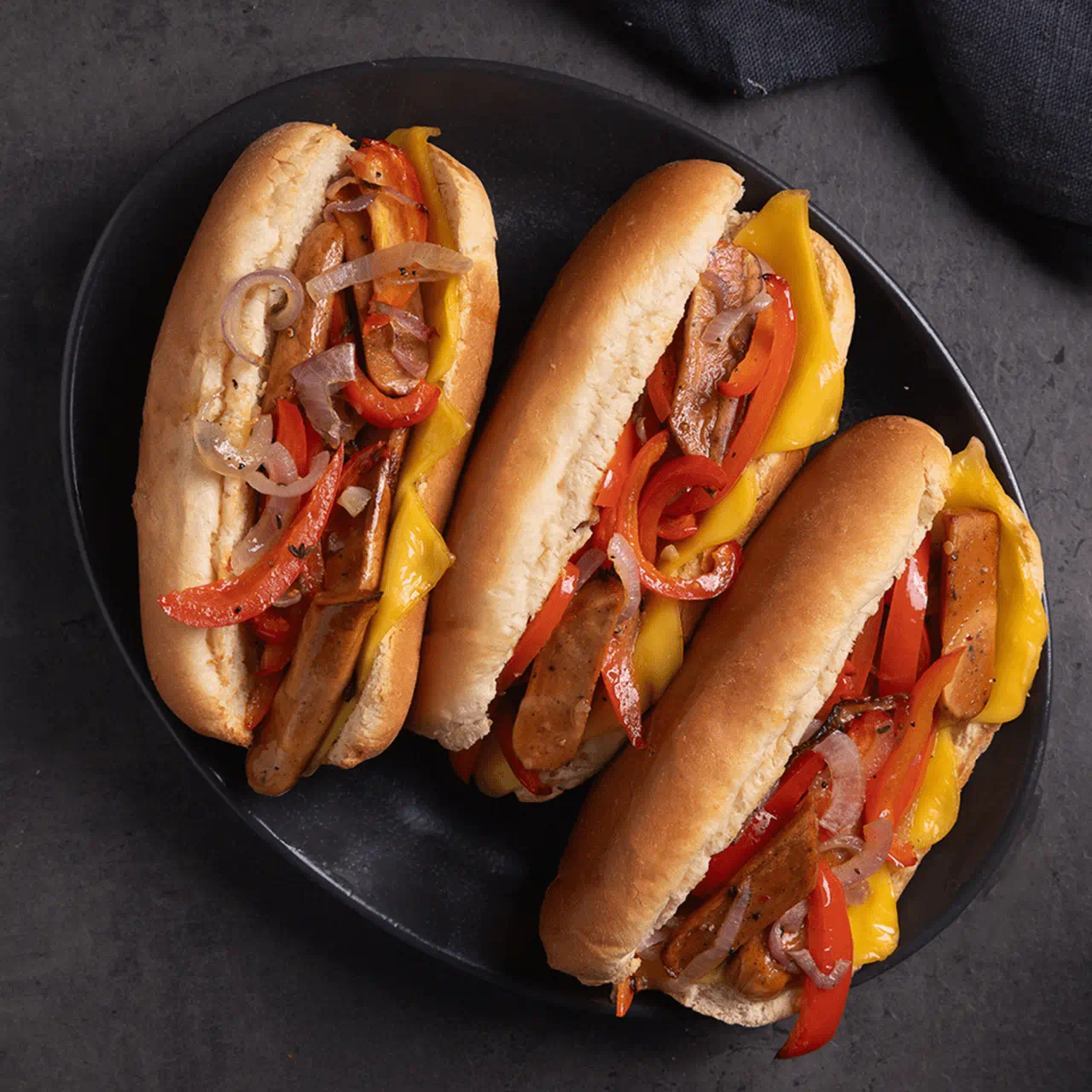 Three sandwich rolls filled with grilled plant based sausages, melted dairy free cheese slices and sautéed red peppers and onions.