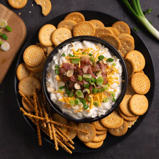 A bowl filled with Million Dollar Dip topped with diary free cheese shreds, bacon, almonds and green onions, served with crackers and pretzel sticks.