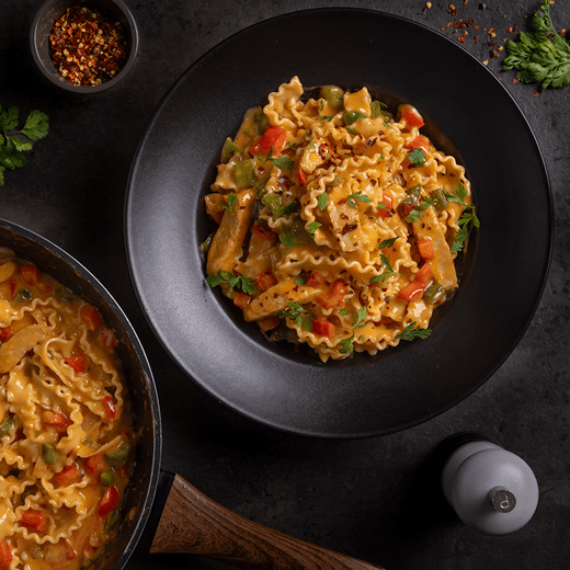 A black bowl filled with pasta, sliced red and green bell peppers and vegan chicken. The dish is sprinkled with fresh herbs and red pepper flakes.