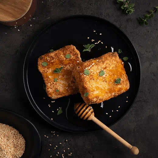 Sesame‑topped vegan Greek saganaki wrapped in crunchy phyllo drizzled with maple syrup and garnished with fresh herbs.