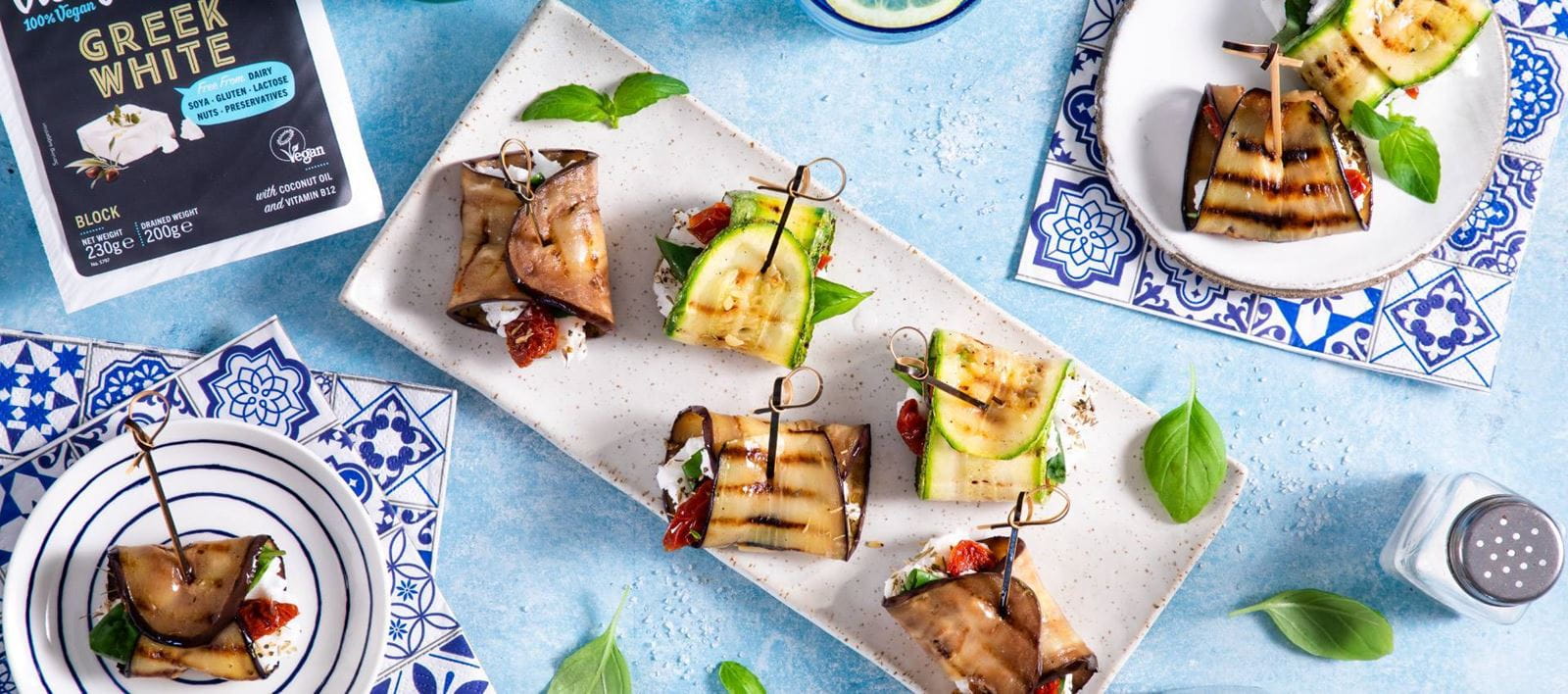 Eggplant and zucchini rolls, each secured with a wooden skewer. Rolls are filled with dairy free cheese in crumbles, basil and sun-dried tomatoes.