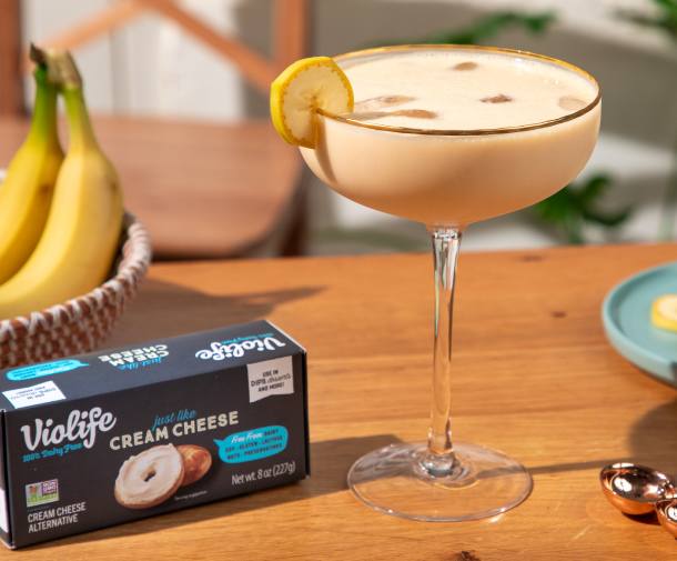 Creamy Banana Split cocktail served in a wide coupe glass garnished with a banana slice. To the left, a box of Violife cream cheese alternative and fresh bananas.
