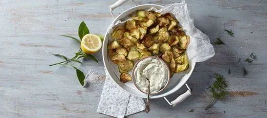 Golden-brown vegan zucchini chips on a round tray. In the center, a small bowl of dairy free creamy dip with herbs and a spoon inside the bowl.