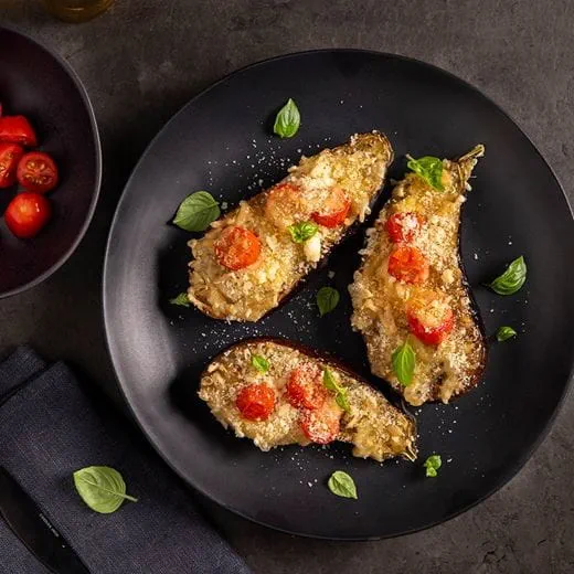 Three halves of baked eggplant topped with a layer of dairy free cheese, breadcrumbs, cherry tomatoes and fresh basil leaves.