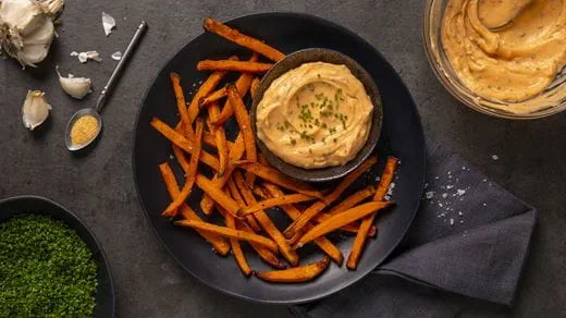 Air fried sweet potato fries served with dairy free creamy dip, topped with chopped chives, surrounded by garlic, seasoning, and fresh herbs.