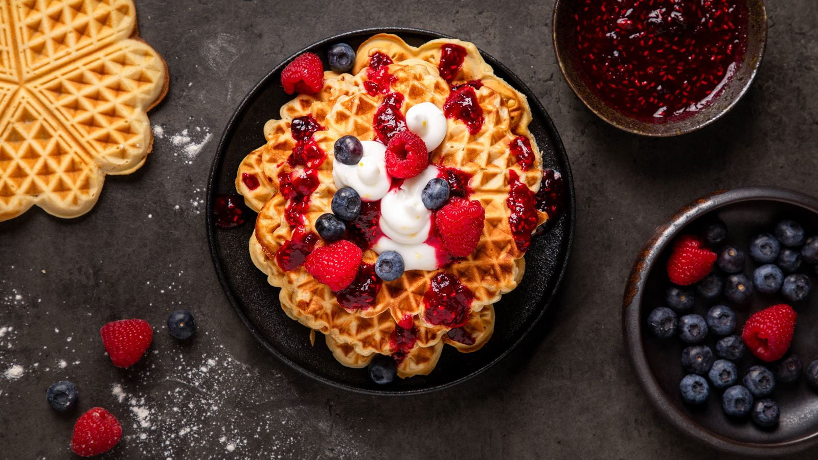 A stack of golden waffles on a dark plate with dairy free cream cheese creamy filing, berry sauce and fresh blueberries and raspberries.
