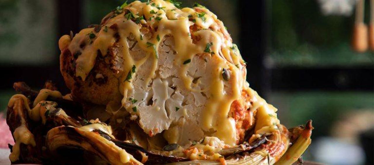 A whole roasted cauliflower cut open to show its interior, topped with Violife dairy free cheesy sauce and sprinkled with chopped herbs.