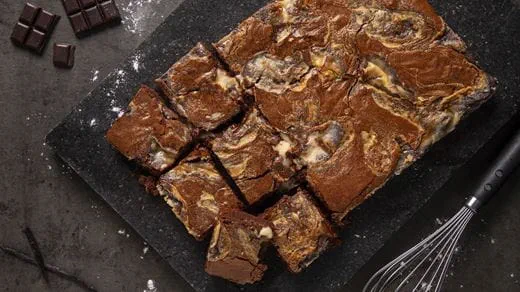 A tray of cheesecake brownies cut into squares, showing a marbled mix of dark chocolate brownie and dairy free creamy swirls.