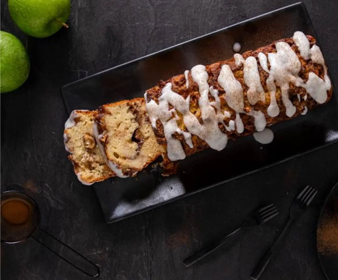 Apple fritter bread drizzled with dairy free frosting on a black plate. Two slices are cut, showing the interior, with apple pieces and cinnamon.