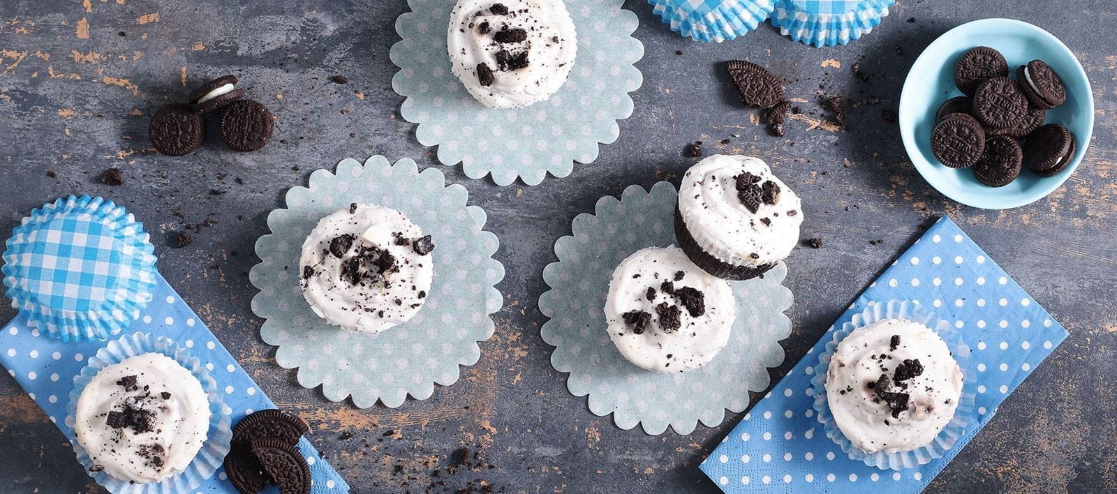 Mini Oreo cheesecake cups arranged on pastel blue paper liners made with dairy free cream cheese, sprinkled with crushed Oreo cookies.