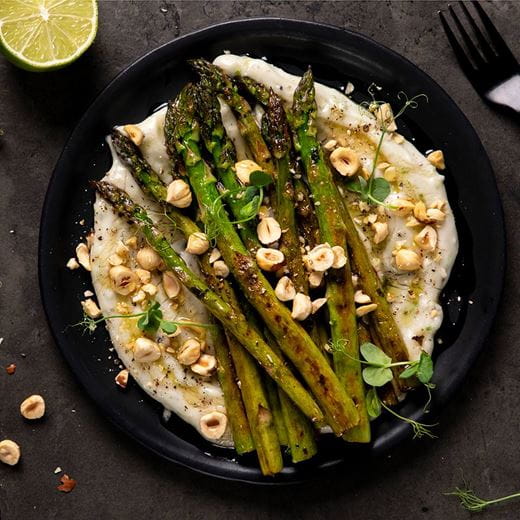 Grilled asparagus spears served on a black plate over a swirl of dairy free cream cheese sauce, topped with chopped hazelnuts and a drizzle of honey.