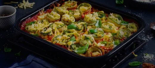 Zucchini rotolo in a rectangular baking dish, thin zucchini slices rolled into spirals and arranged upright in rows, filled with peas, dairy free cheese and basil.
