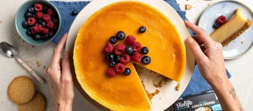 A round dairy free cheesecake with a smooth golden top in a white plate, topped with fresh raspberries and blueberries.
