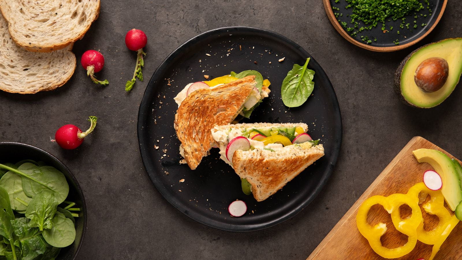 Toasted rye bread sandwich cut in half on a black plate, filled with sliced radishes, yellow bell pepper, spinach leaves and dairy free spread.