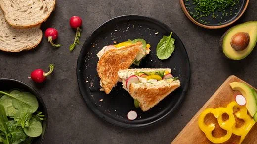Toasted rye bread sandwich cut in half on a black plate, filled with sliced radishes, yellow bell pepper, spinach leaves and dairy free spread.