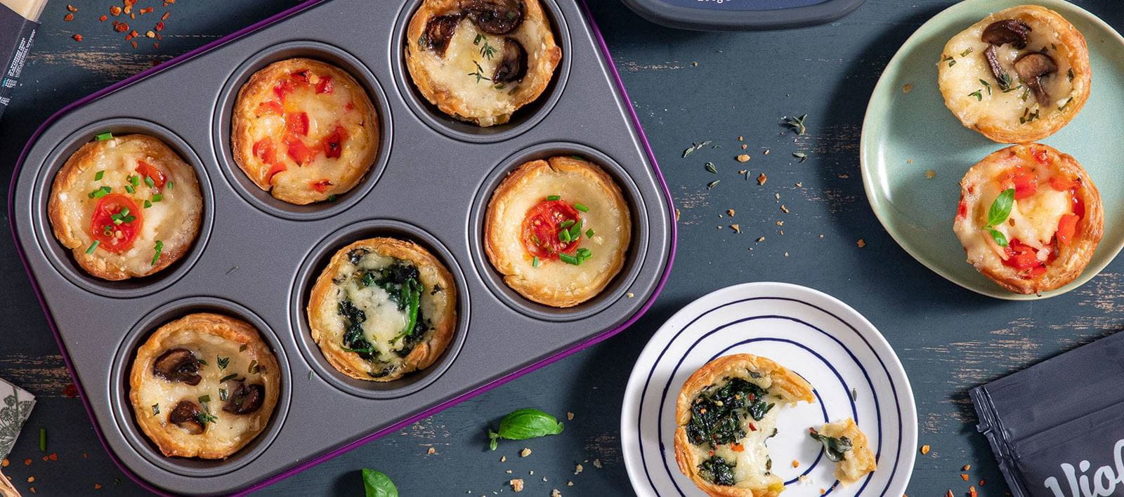 A muffin tin filled with small vegan savory tarts with various toppings, including melted cheese, cherry tomatoes, mushrooms and spinach.