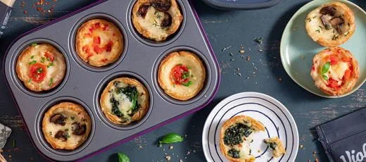 A muffin tin filled with small vegan savory tarts with various toppings, including melted cheese, cherry tomatoes, mushrooms and spinach.