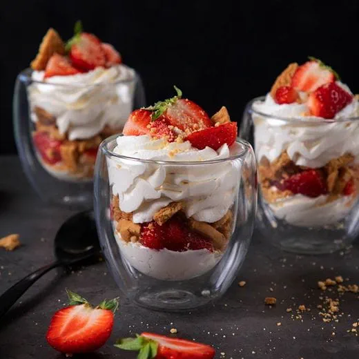 Three glass cups filled with layers of whipped dairy free cream cheese, fresh strawberry pieces and crumbled cookies.