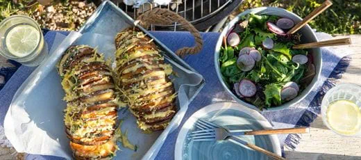 Hasselback sweet potatoes in a metal tray lined with parchment, each sliced thinly and topped with melted dairy free cheese, herbs, and seasoning.