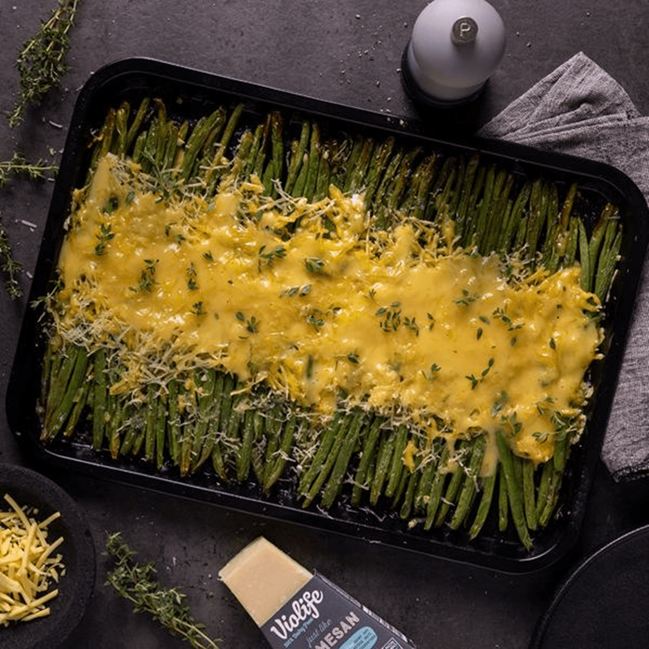 A baking tray filled with roasted green beans arranged neatly in rows, topped with a layer of melted dairy free cheese and sprinkled with fresh herbs.