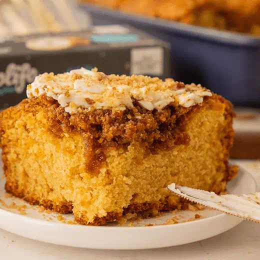 A thick slice of golden coffee cake topped with a layer of crumbly streusel and swirls of cream cheese frosting, on a white plate.