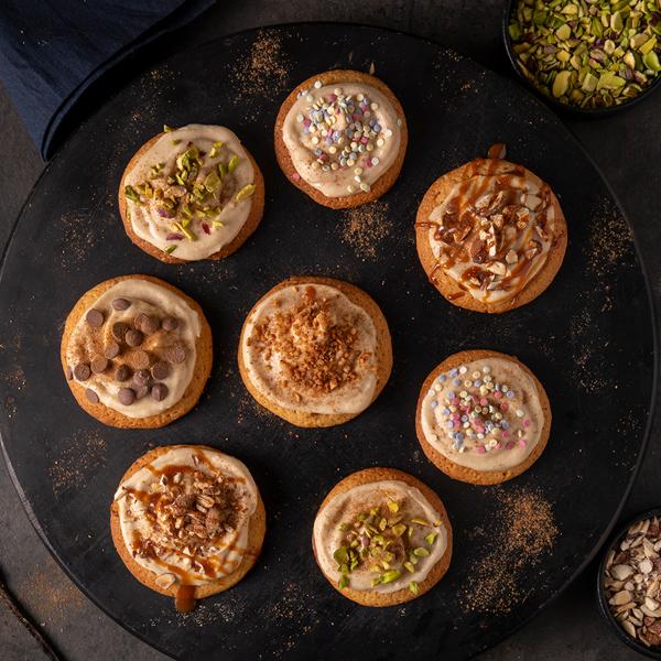 Eight Crumbl Cookies each topped with dairy free caramel frosting and different mix‑ins such as chopped pistachios, colorful sprinkles and chocolate chips.