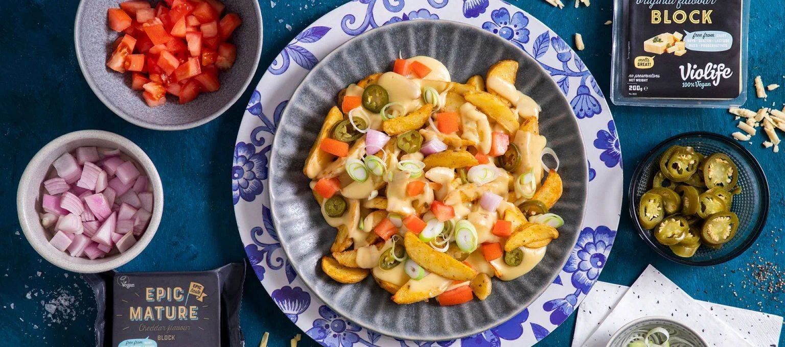 A bowl filled with seasoned potato wedges topped with dairy free cheese sauce, diced tomatoes, red onions, sliced green onions and jalapeño rounds.