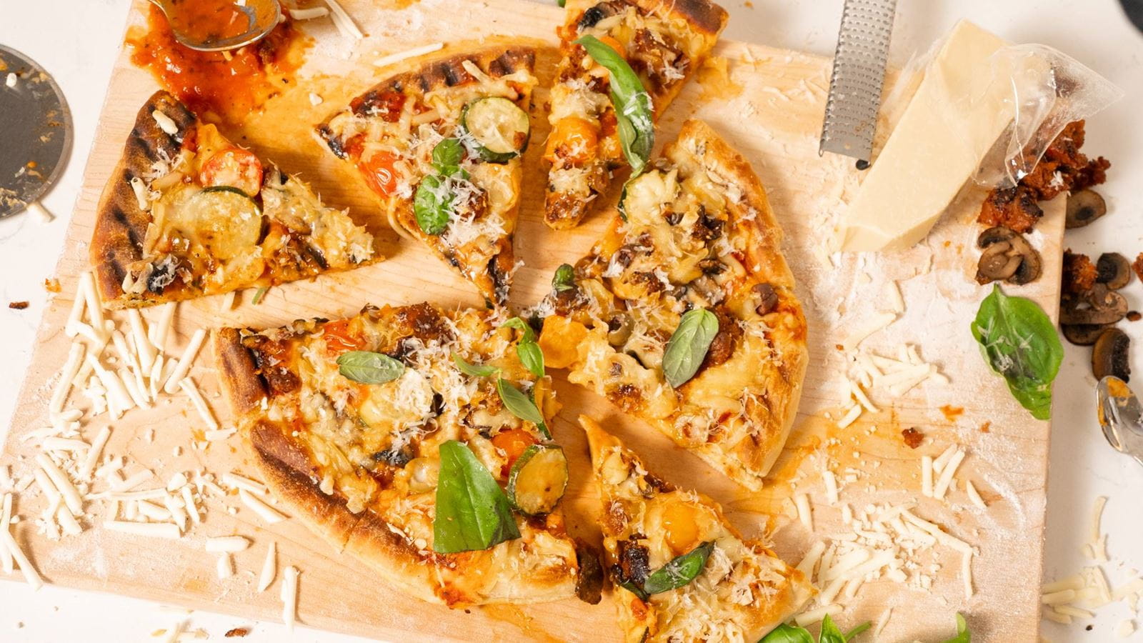 A sliced grilled pizza on a wooden board, topped with dairy free cheese, vegetables like grilled eggplant and zucchini and fresh basil leaves.
