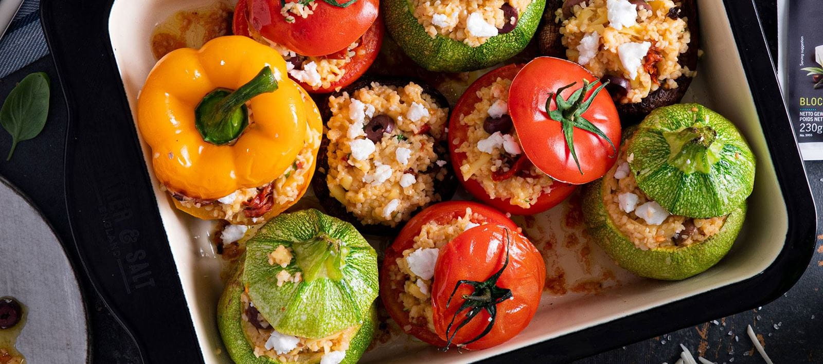 Stuffed vegetables in a baking dish, round zucchini, tomatoes and a yellow bell pepper filled with rice, dairy free cheese crumbles and olive pieces.