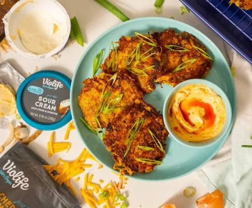 A plate of crispy turmeric latkes topped with sliced green onions, served with a bowl of dairy free sour cream drizzled with chili crisp.