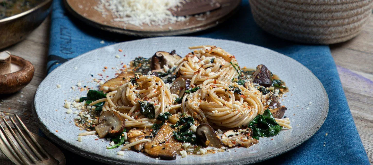 A plate of spaghetti formed into small nests, mixed with sautéed mushrooms, spinach and dairy free cheese, served on a grey dish.