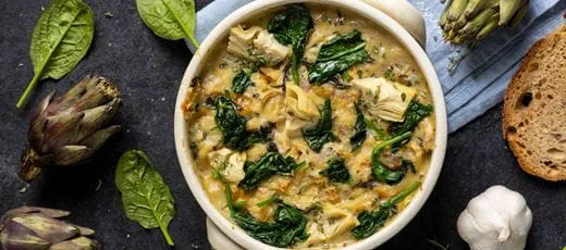 A bowl of creamy spinach and artichoke dip made with wilted fresh spinach, artichoke hearts, dairy free cheese, onion and garlic, served with slices of bread.